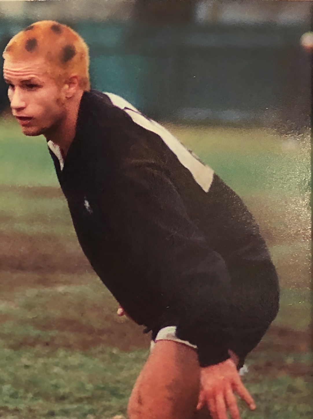 Nick Taylor crouched on a rugby field in a black jersey, short hair bleached in blonde spots.